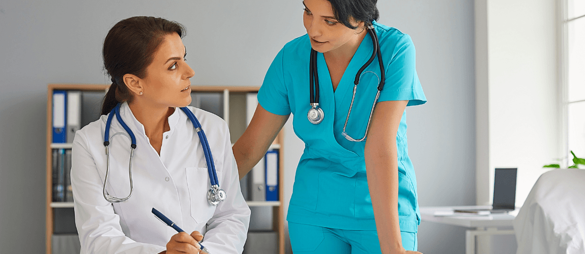 Eine Ärztin sitzt an einem Tisch und sieht eine dahinter stehende Versorgungsassistentin an. 