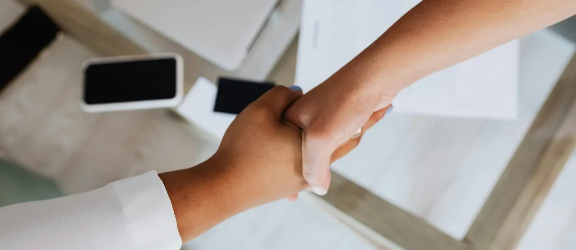 Zwei Hände halten sich, im Hintergrund sind Unterlagen und ein Handy auf einem Tisch zu sehen.