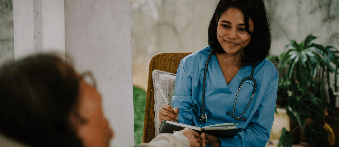 Eine Pflegefachkraft sitzt einer Pflegeperson gegenüber, die im Bett liegt.