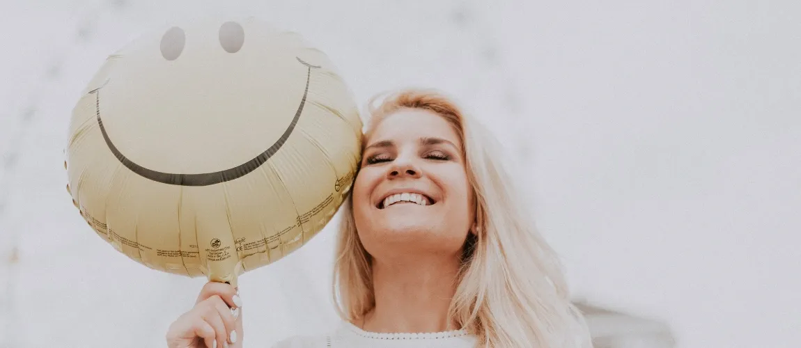 Eine Frau hält einen Ballon mit einem Lächeln darauf neben ihr Gesicht und lacht.