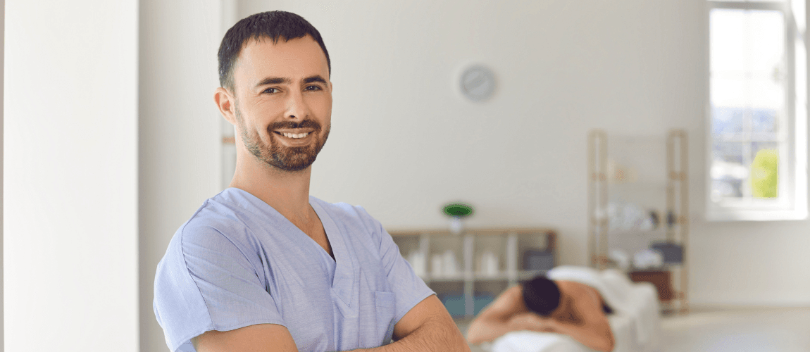 Ein Mann lächelt in die Kamera, im Hintergrund liegt eine Frau auf einer Behandlungsliege. 