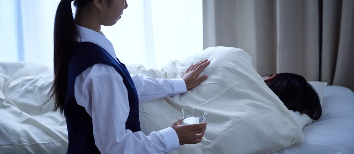 Ein Mann liegt im Bett. Eine Frau steht daneben und legt ihm die Hand auf dem Rücken.