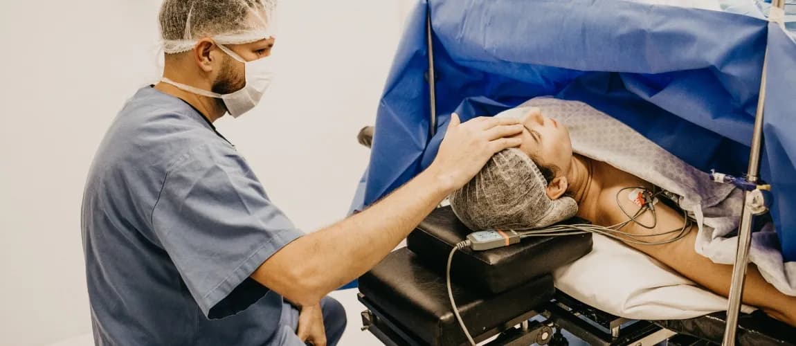 Eine Frau liegt auf einem Operationstisch, hinter ihr sitzt ein Narkosearzt und legt der Frau die Hand auf die Stirn.