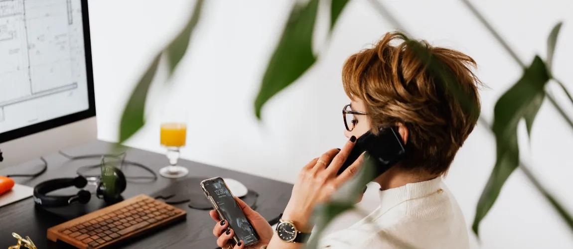 Eine Frau telefoniert am Schreibtisch und blickt gleichzeitig auf ihr Smartphone.