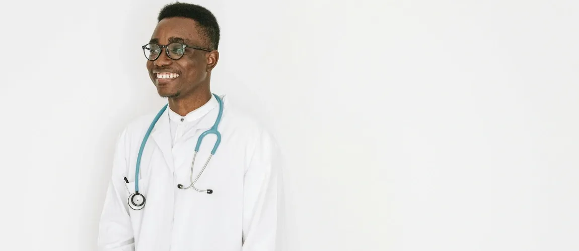 Ein Pflegefachmann steht in Berufskleidung vor der Kamera und lächelt.