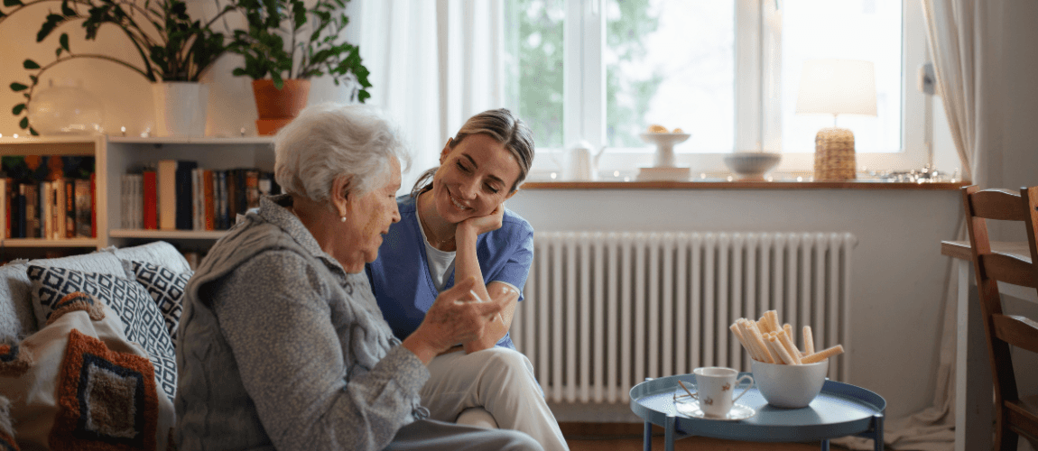 Eine Pflegehelferin sitzt mit einer Patientin auf dem Sofa.