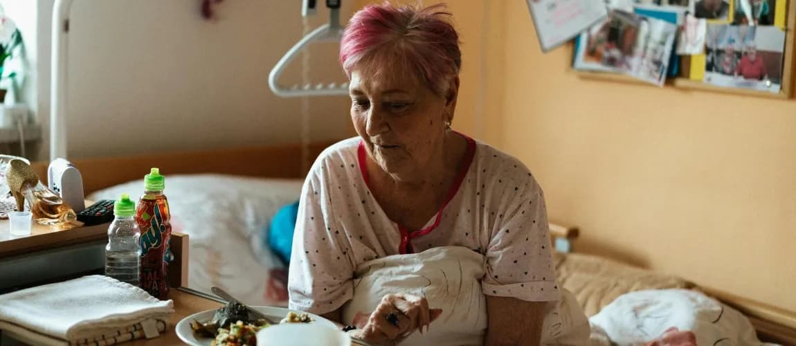 Eine ältere Frau sitzt im Pflegebett und schaut zögerlich auf einen Teller mit einer Speise.
