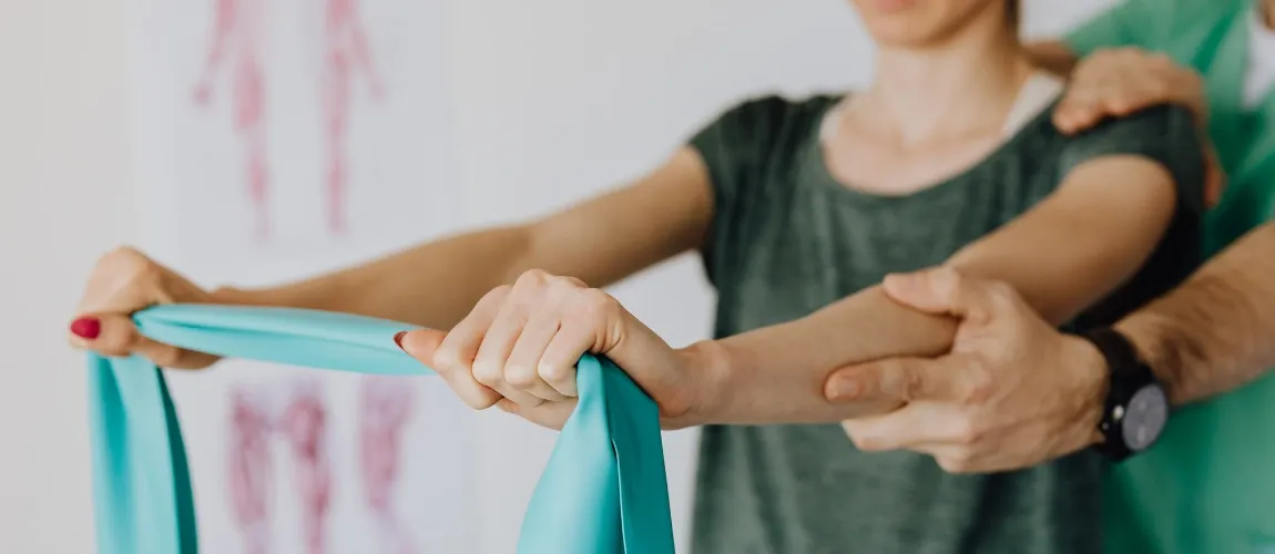 Eine Frau hält ein Trainingsband in den Händen, ein Physiotherapeut steht dahinter und stützt den Arm.