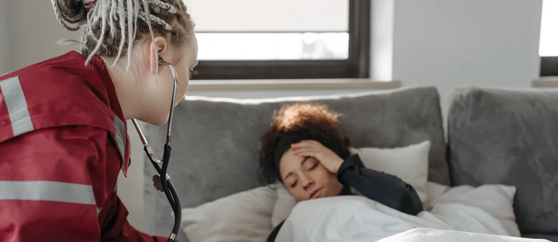 Eine Frau liegt im Bett und fasst sich mit einer Hand an den Kopf, eine Rettungssanitäterin ist dabei.