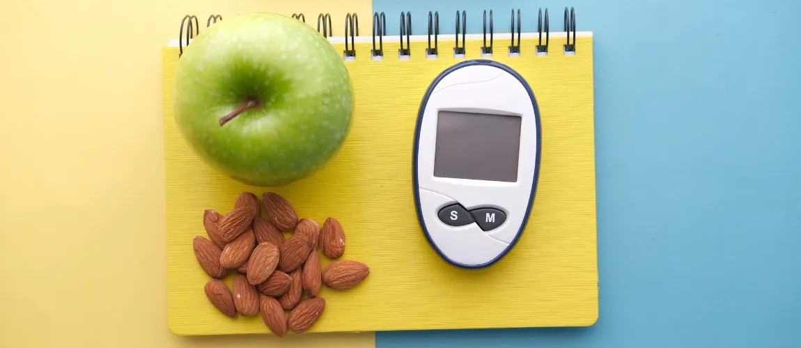 Ein Apfel und Mandeln liegen auf einem Notizbuch, daneben ein Blutzuckermessgerät.
