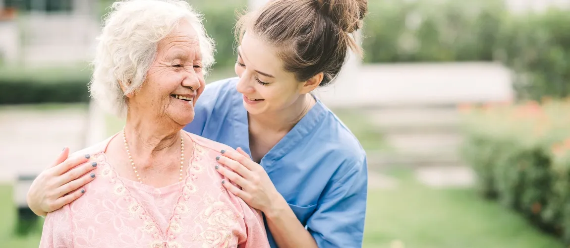 Es ist eine Pflegerin und eine Patientin beim Spaziergang im Garten zu sehen. 