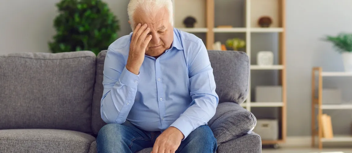 Ein Mann sitzt auf einem Sofa und fasst sich mit einer Hand an die Stirn.