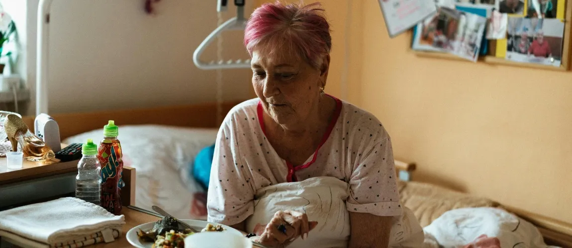 Eine ältere Frau sitzt im Pflegebett und schaut zögerlich auf einen Teller mit einer Speise.