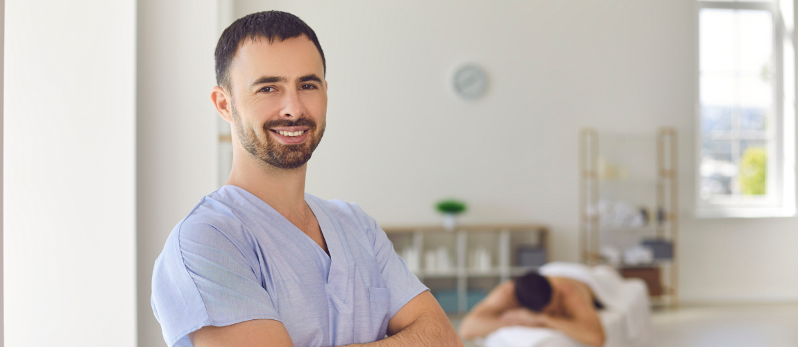 Ein Mann lächelt in die Kamera, im Hintergrund liegt eine Frau auf einer Behandlungsliege. 