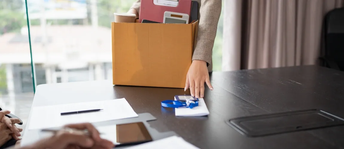 Eine Frau schiebt einen Zettel über einen Tisch, vor ihr steht ein Karton mit Habseligkeiten. 