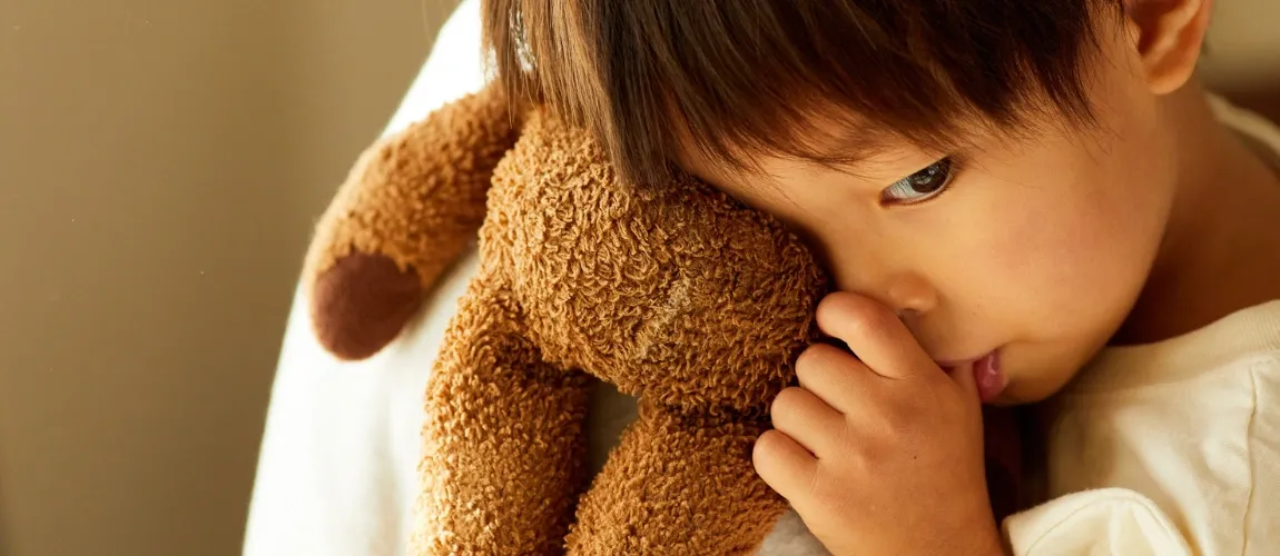 Ein Kleinkind umarmt einen Teddybären und lutscht am Daumen.