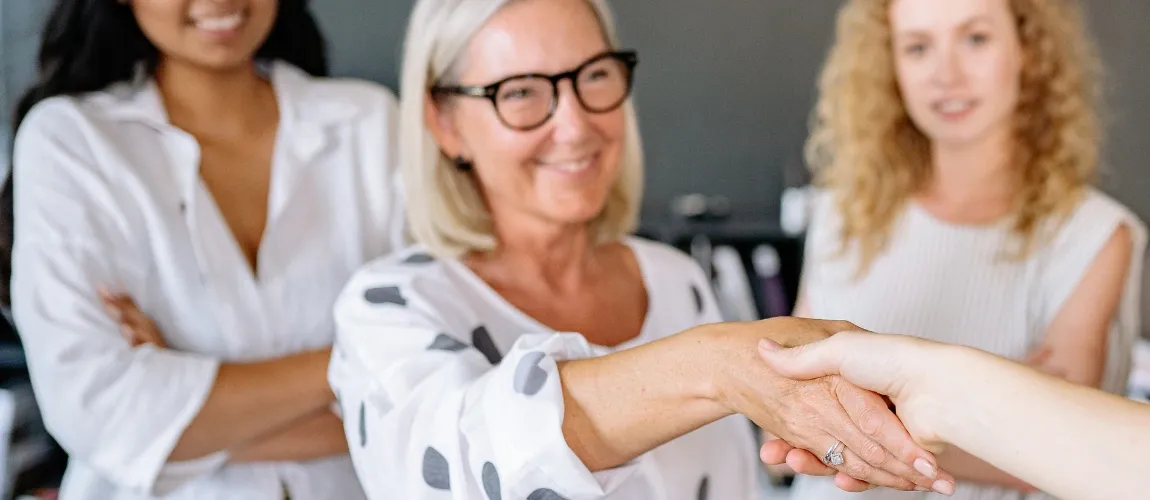 Eine Frau lächelt und gibt einer Person die Hand, im Hintergrund stehen zwei Frauen.